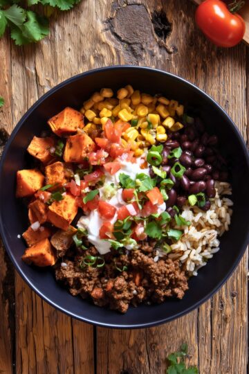 Overhead view of a Beef Burrito Sweet Potato Bowl in a matte black bowl with roasted sweet potato cubes, seasoned ground beef, black beans, rice, corn, diced tomatoes, sour cream, green onions, and cilantro. Served on a rustic wooden table.