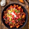 Overhead view of a Beef Enchilada Sweet Potato Bowl with roasted sweet potatoes, seasoned ground beef, black beans, shredded cheese, tomatoes, red onion, cilantro, and sour cream in a black bowl on a rustic wood table.