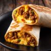 Two Beef Taco Breakfast Burrito halves stacked on a matte black plate. Lightly toasted flour tortillas reveal seasoned ground beef, fluffy scrambled eggs, melted cheddar cheese, and green onions. Dark rustic wood background.
