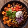 Close-up overhead view of a Beef Taco Sweet Potato Bowl filled with seasoned ground beef, roasted sweet potatoes, black beans, sweet corn, diced tomatoes, chopped red onion, shredded cheese, sour cream, and fresh cilantro in a matte black bowl on a rustic wooden table.