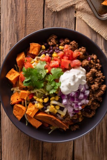 Close-up overhead view of a Beef Taco Sweet Potato Bowl filled with seasoned ground beef, roasted sweet potatoes, black beans, sweet corn, diced tomatoes, chopped red onion, shredded cheese, sour cream, and fresh cilantro in a matte black bowl on a rustic wooden table.