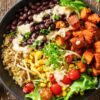 Close-up of a black bean sweet potato protein bowl with seasoned roasted sweet potato cubes, black beans, fluffy quinoa, sweet corn, cherry tomatoes, lettuce, shredded cheese, fresh herbs, and creamy white dressing in a matte black bowl on rustic wood.