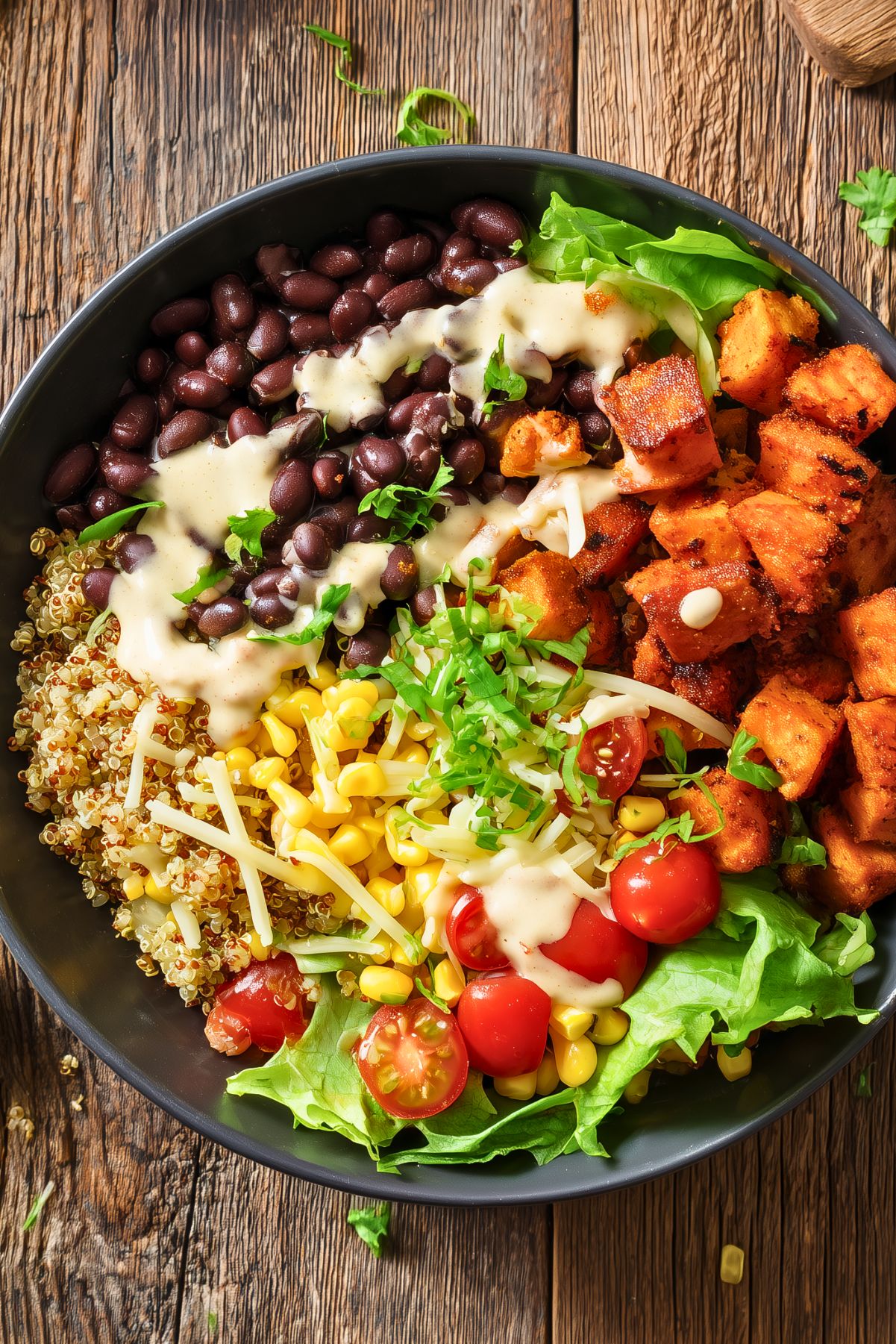 Close-up of a black bean sweet potato protein bowl with seasoned roasted sweet potato cubes, black beans, fluffy quinoa, sweet corn, cherry tomatoes, lettuce, shredded cheese, fresh herbs, and creamy white dressing in a matte black bowl on rustic wood.