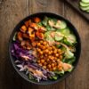 Overhead view of a Buffalo chickpea sweet potato bowl with roasted sweet potatoes, seasoned chickpeas, romaine, cucumber, red cabbage, and creamy Buffalo dressing on a rustic wood table.