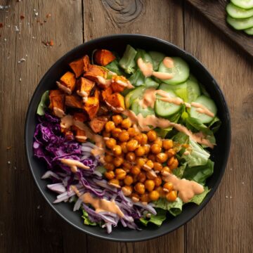 Overhead view of a Buffalo chickpea sweet potato bowl with roasted sweet potatoes, seasoned chickpeas, romaine, cucumber, red cabbage, and creamy Buffalo dressing on a rustic wood table.