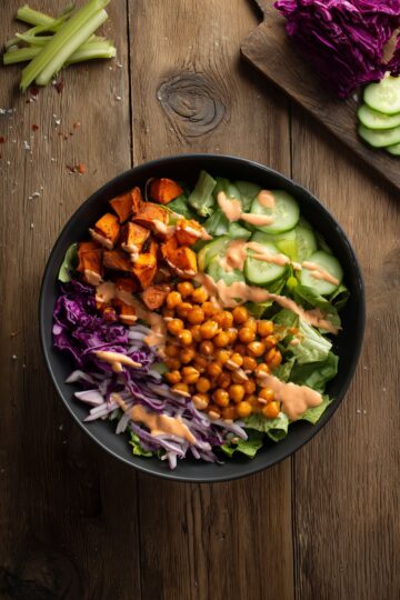 Overhead view of a Buffalo chickpea sweet potato bowl with roasted sweet potatoes, seasoned chickpeas, romaine, cucumber, red cabbage, and creamy Buffalo dressing on a rustic wood table.