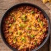 Overhead view of Cheesy Sloppy Joe Party Dip in a round black baking dish. Seasoned ground beef in a rich tomato sauce is covered with melted cheddar cheese and chopped green onions. Tortilla chips sit nearby on a rustic wooden table.