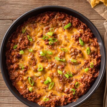Overhead view of Cheesy Sloppy Joe Party Dip in a round black baking dish. Seasoned ground beef in a rich tomato sauce is covered with melted cheddar cheese and chopped green onions. Tortilla chips sit nearby on a rustic wooden table.