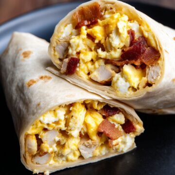 Close-up of two stacked Chicken Bacon Ranch Breakfast Burrito halves on a black plate. Golden scrambled eggs, chunks of chicken breast, crispy bacon, and melted cheddar cheese fill the lightly toasted flour tortillas.