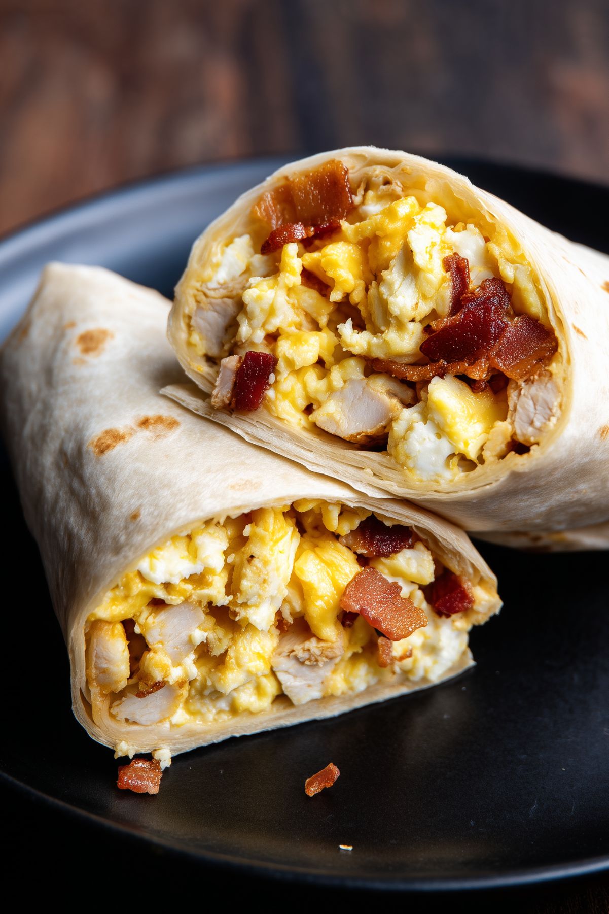 Close-up of two stacked Chicken Bacon Ranch Breakfast Burrito halves on a black plate. Golden scrambled eggs, chunks of chicken breast, crispy bacon, and melted cheddar cheese fill the lightly toasted flour tortillas.