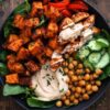 Close-up of a Chicken and Hummus Sweet Potato Bowl with golden roasted sweet potatoes, seasoned chicken drizzled with creamy white sauce, chickpeas, sliced cucumbers, red bell peppers, fresh spinach, and a generous scoop of hummus in a black bowl.