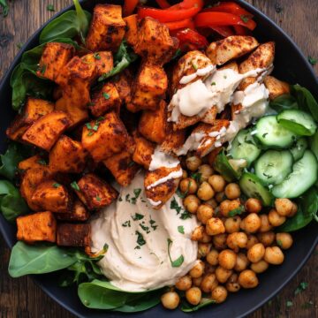 Close-up of a Chicken and Hummus Sweet Potato Bowl with golden roasted sweet potatoes, seasoned chicken drizzled with creamy white sauce, chickpeas, sliced cucumbers, red bell peppers, fresh spinach, and a generous scoop of hummus in a black bowl.