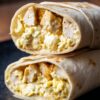Close-up of two stacked chicken and waffle breakfast burrito halves filled with scrambled eggs, seasoned chicken, melted cheese, and waffle pieces on a black plate.