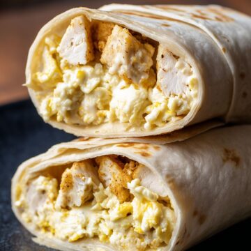 Close-up of two stacked chicken and waffle breakfast burrito halves filled with scrambled eggs, seasoned chicken, melted cheese, and waffle pieces on a black plate.