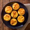 Seven homemade Chipotle Cheddar Egg Bites arranged on a matte black plate. The golden yellow egg bites have lightly browned, cheesy tops with visible chipotle seasoning. A beige napkin rests beneath the plate on a rustic wooden table.