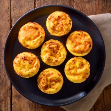 Seven homemade Chipotle Cheddar Egg Bites arranged on a matte black plate. The golden yellow egg bites have lightly browned, cheesy tops with visible chipotle seasoning. A beige napkin rests beneath the plate on a rustic wooden table.