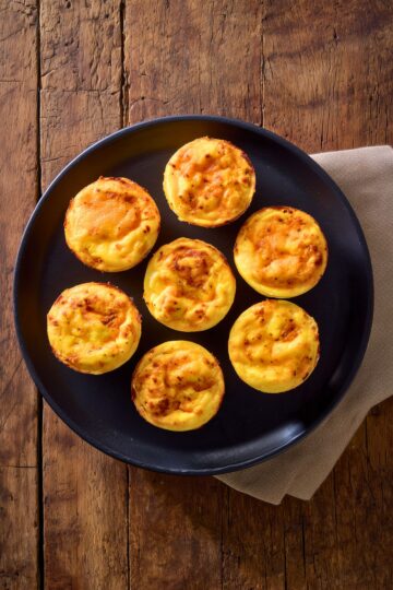 Seven homemade Chipotle Cheddar Egg Bites arranged on a matte black plate. The golden yellow egg bites have lightly browned, cheesy tops with visible chipotle seasoning. A beige napkin rests beneath the plate on a rustic wooden table.