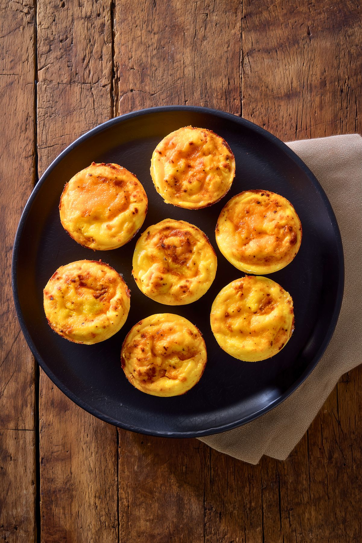Seven homemade Chipotle Cheddar Egg Bites arranged on a matte black plate. The golden yellow egg bites have lightly browned, cheesy tops with visible chipotle seasoning. A beige napkin rests beneath the plate on a rustic wooden table.