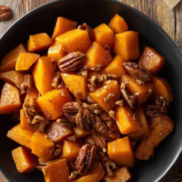 Close-up of cinnamon roasted butternut squash with caramelized edges and toasted pecans in a matte black bowl on a rustic wood table.