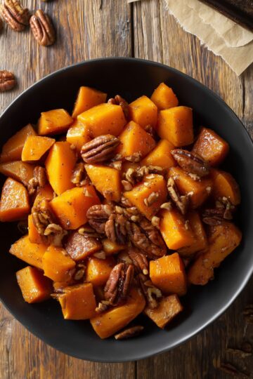 Close-up of cinnamon roasted butternut squash with caramelized edges and toasted pecans in a matte black bowl on a rustic wood table.