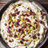 Close overhead view of Cranberry Pistachio Cream Cheese Dip in a round black bowl. Thick, creamy dip with swirls of cream cheese, dried red cranberries, and chopped green pistachios on a rustic wooden table.