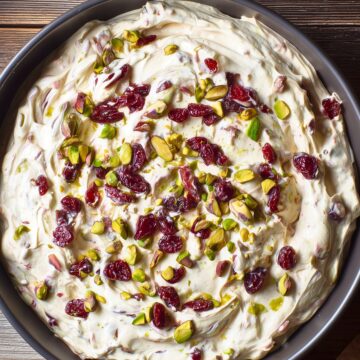 Close overhead view of Cranberry Pistachio Cream Cheese Dip in a round black bowl. Thick, creamy dip with swirls of cream cheese, dried red cranberries, and chopped green pistachios on a rustic wooden table.