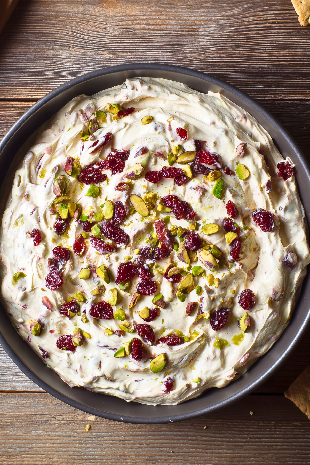 Close overhead view of Cranberry Pistachio Cream Cheese Dip in a round black bowl. Thick, creamy dip with swirls of cream cheese, dried red cranberries, and chopped green pistachios on a rustic wooden table.