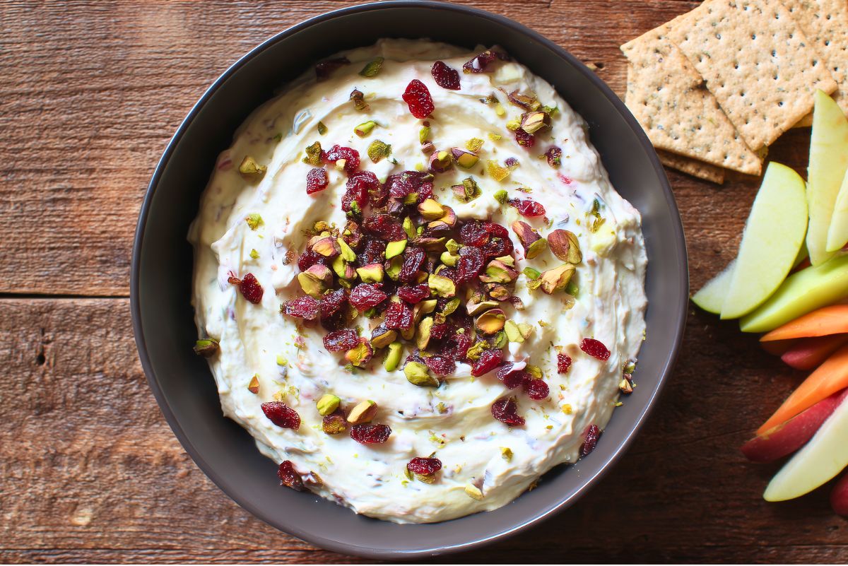 Overhead view of Cranberry Pistachio Cream Cheese Dip in a black bowl, topped with dried cranberries and chopped pistachios. Served on a rustic wooden table with crackers, sliced apples, and carrot sticks alongside.