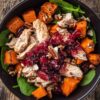 Overhead close-up of a cranberry turkey sweet potato bowl with tender turkey pieces, roasted orange sweet potato cubes, glossy cranberry sauce, fresh baby spinach, and crunchy pecans in a matte black bowl on a rustic wooden table.