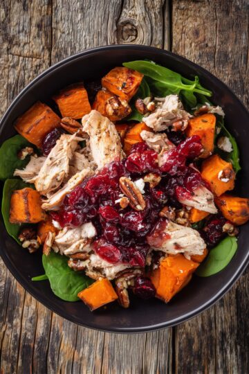 Overhead close-up of a cranberry turkey sweet potato bowl with tender turkey pieces, roasted orange sweet potato cubes, glossy cranberry sauce, fresh baby spinach, and crunchy pecans in a matte black bowl on a rustic wooden table.