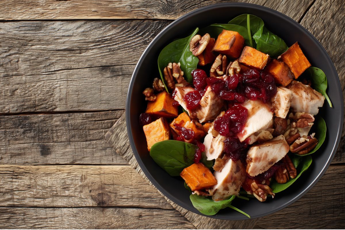 Overhead view of a cranberry turkey sweet potato bowl with sliced turkey breast topped with cranberry sauce, roasted sweet potato cubes, leafy spinach, and pecans. Served in a matte black bowl on rustic wood with open space to the left.
