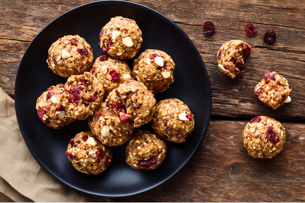 Homemade Cranberry White Chocolate Protein Balls piled on a matte black plate on a rustic wooden table. Oatmeal snack balls studded with dried cranberries and white chocolate chips, with additional protein balls and cranberries scattered nearby.