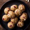 Overhead close-up of Cranberry White Chocolate Protein Balls arranged on a black plate. Golden oat and almond butter protein bites contain red dried cranberries and creamy white chocolate chips, with scattered ingredients on a dark rustic wooden background.