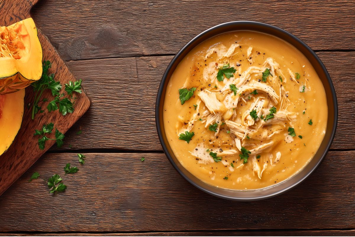 Overhead view of Creamy Roasted Acorn Squash Chicken Soup in a black bowl, topped with shredded chicken, chopped parsley, and black pepper. Sliced acorn squash and fresh parsley sit on a wooden cutting board beside the bowl on a dark rustic wood table.
