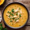 Creamy Roasted Acorn Squash Chicken Soup in a large black bowl, featuring a thick, golden-orange broth filled with shredded chicken and garnished with fresh parsley and black pepper. A wedge of acorn squash and fresh parsley rest nearby on a rustic wooden table.