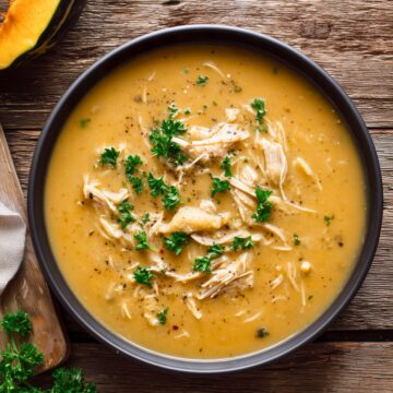 Creamy Roasted Acorn Squash Chicken Soup in a large black bowl, featuring a thick, golden-orange broth filled with shredded chicken and garnished with fresh parsley and black pepper. A wedge of acorn squash and fresh parsley rest nearby on a rustic wooden table.