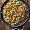 Overhead view of creamy green bean casserole with tender green beans, sliced mushrooms, and crispy golden fried onions in a matte black bowl on a rustic wooden cutting board with a beige napkin.