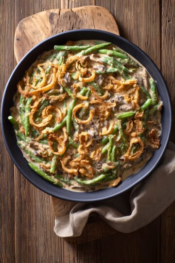 Overhead view of creamy green bean casserole with tender green beans, sliced mushrooms, and crispy golden fried onions in a matte black bowl on a rustic wooden cutting board with a beige napkin.