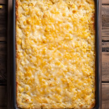Overhead view of homemade creamy corn casserole baked in a rectangular black baking dish. The golden brown casserole has a creamy texture, visible sweet corn kernels, and melted cheddar cheese across the top, resting on rustic dark wood.