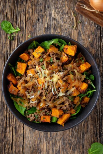 Overhead view of a French Onion Beef Sweet Potato Bowl filled with seasoned ground beef, golden caramelized onions, roasted sweet potato cubes, fresh spinach, and shredded cheese. Served in a matte black bowl on a rustic wooden table.