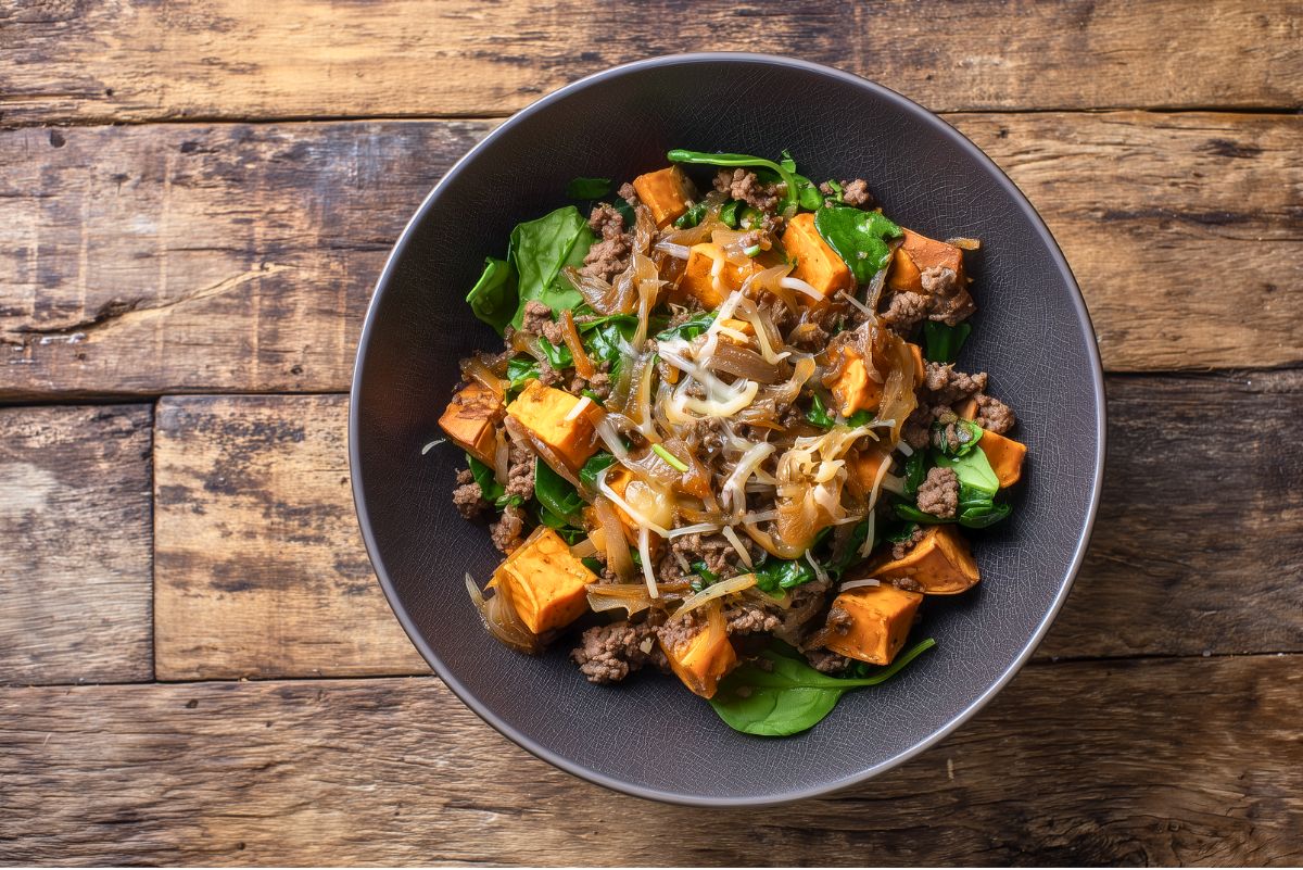 Overhead view of a French Onion Beef Sweet Potato Bowl with browned ground beef, caramelized onions, roasted sweet potato cubes, fresh spinach, and melted shredded cheese. Served in a round black bowl centered on a rustic wooden table.