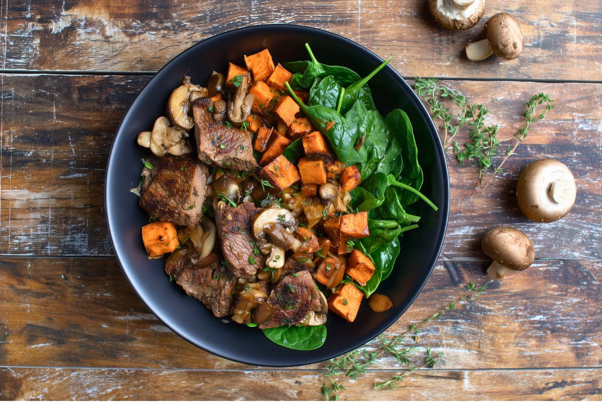 Overhead view of a Garlic Mushroom Steak Sweet Potato Bowl with seared steak bites, garlic mushrooms, roasted sweet potato cubes, and fresh baby spinach. Served in a black bowl on a rustic wooden table with fresh thyme and whole mushrooms nearby.
