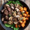 Close-up of a Garlic Mushroom Steak Sweet Potato Bowl filled with juicy browned steak bites, sautéed sliced mushrooms, golden roasted sweet potato cubes, and fresh spinach. Garnished with herbs and served in a matte black bowl on a rustic wooden table.