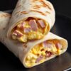 Close-up of two ham egg and cheese breakfast burrito halves stacked on a matte black plate. Lightly toasted flour tortillas reveal a filling of fluffy scrambled eggs, diced ham, and melted cheddar cheese.