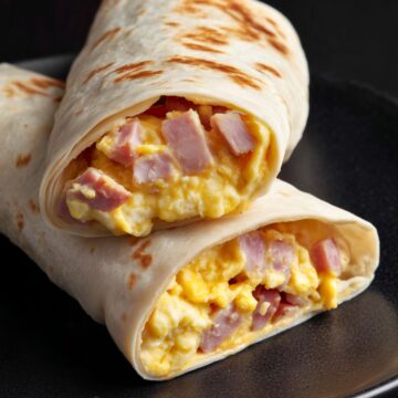 Close-up of two ham egg and cheese breakfast burrito halves stacked on a matte black plate. Lightly toasted flour tortillas reveal a filling of fluffy scrambled eggs, diced ham, and melted cheddar cheese.