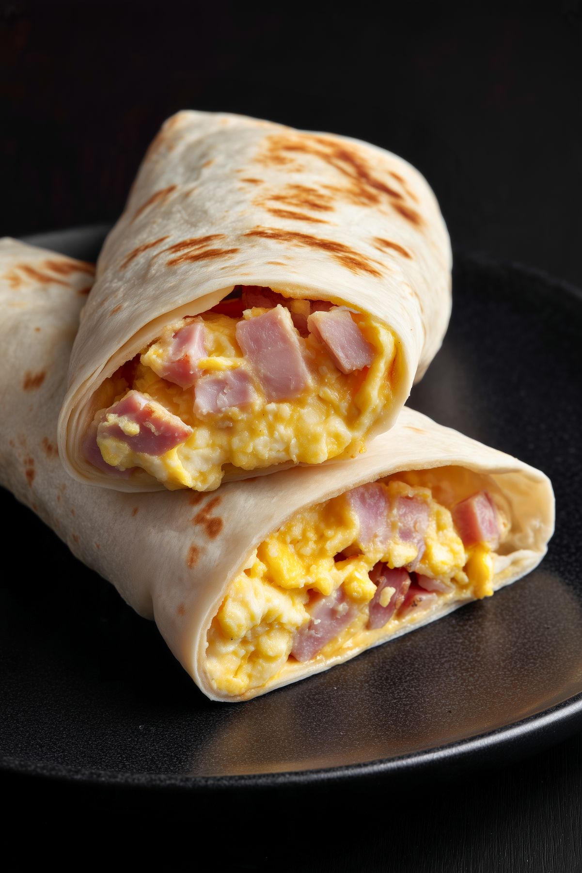 Close-up of two ham egg and cheese breakfast burrito halves stacked on a matte black plate. Lightly toasted flour tortillas reveal a filling of fluffy scrambled eggs, diced ham, and melted cheddar cheese.
