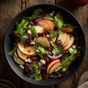 Overhead view of Harvest Apple Salad with Cider Dressing in a matte black bowl. Fresh mixed greens are topped with sliced red apples, dried cranberries, pecans, crumbled feta cheese, red onion, and golden cider dressing on a rustic wooden table.