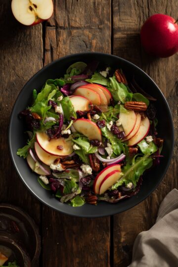 Overhead view of Harvest Apple Salad with Cider Dressing in a matte black bowl. Fresh mixed greens are topped with sliced red apples, dried cranberries, pecans, crumbled feta cheese, red onion, and golden cider dressing on a rustic wooden table.