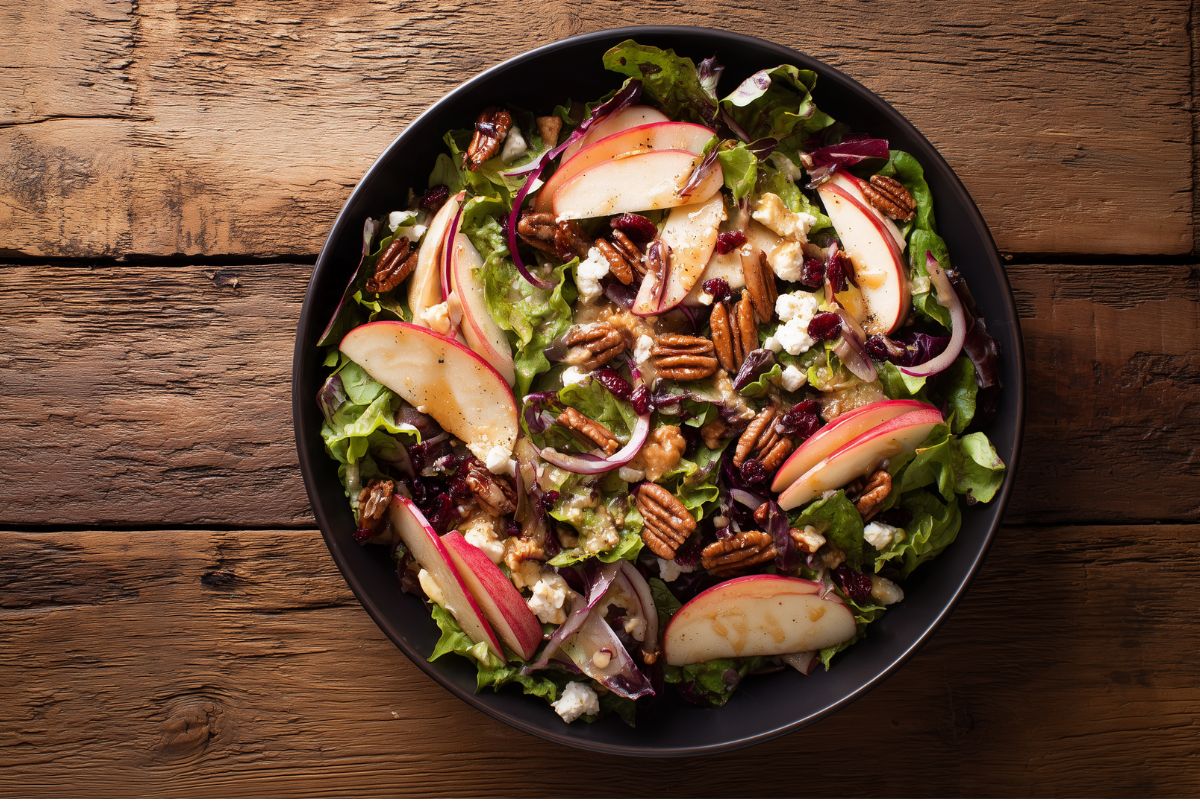 Harvest Apple Salad with Cider Dressing served in a round black bowl on rustic wood. Crisp red apple slices, mixed greens, dried cranberries, toasted pecans, feta cheese, and thin red onion slices are coated with golden apple cider dressing.