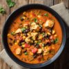 A large black bowl of high protein chicken enchilada soup filled with tender chicken pieces, black beans, sweet corn, diced tomatoes, and green peppers in a seasoned orange-red broth. Fresh cilantro is sprinkled on top, with a beige napkin underneath on a rustic wooden table.